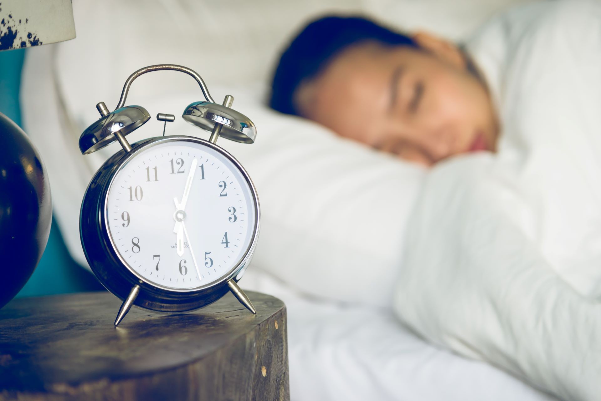 An alarm clock on a bedside table, with an off-focus sleeping woman in the background