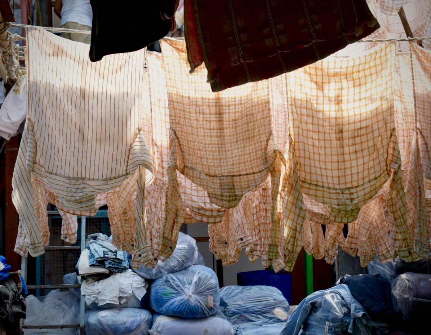 A dozen of white and beige collar shirts, hung to dry on clothes lines. In the background, we can see blue plastic bags that seem to be filled with pieces of clothing. 
