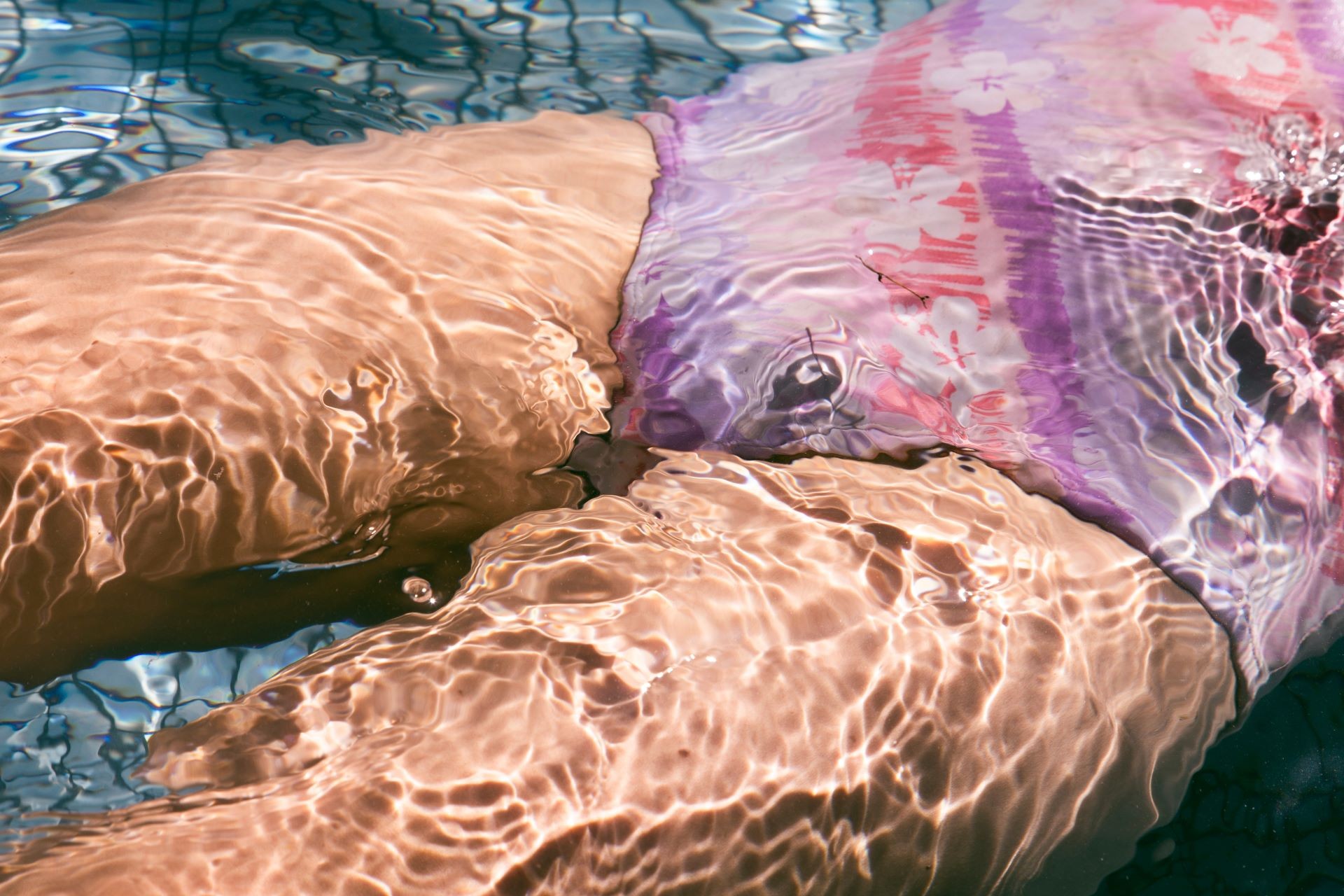The bottom half of a girl's body, under water. The girl is wearing a pink and purple swimsuit. The sun is shining through the water. The bottom of the pool is blue.