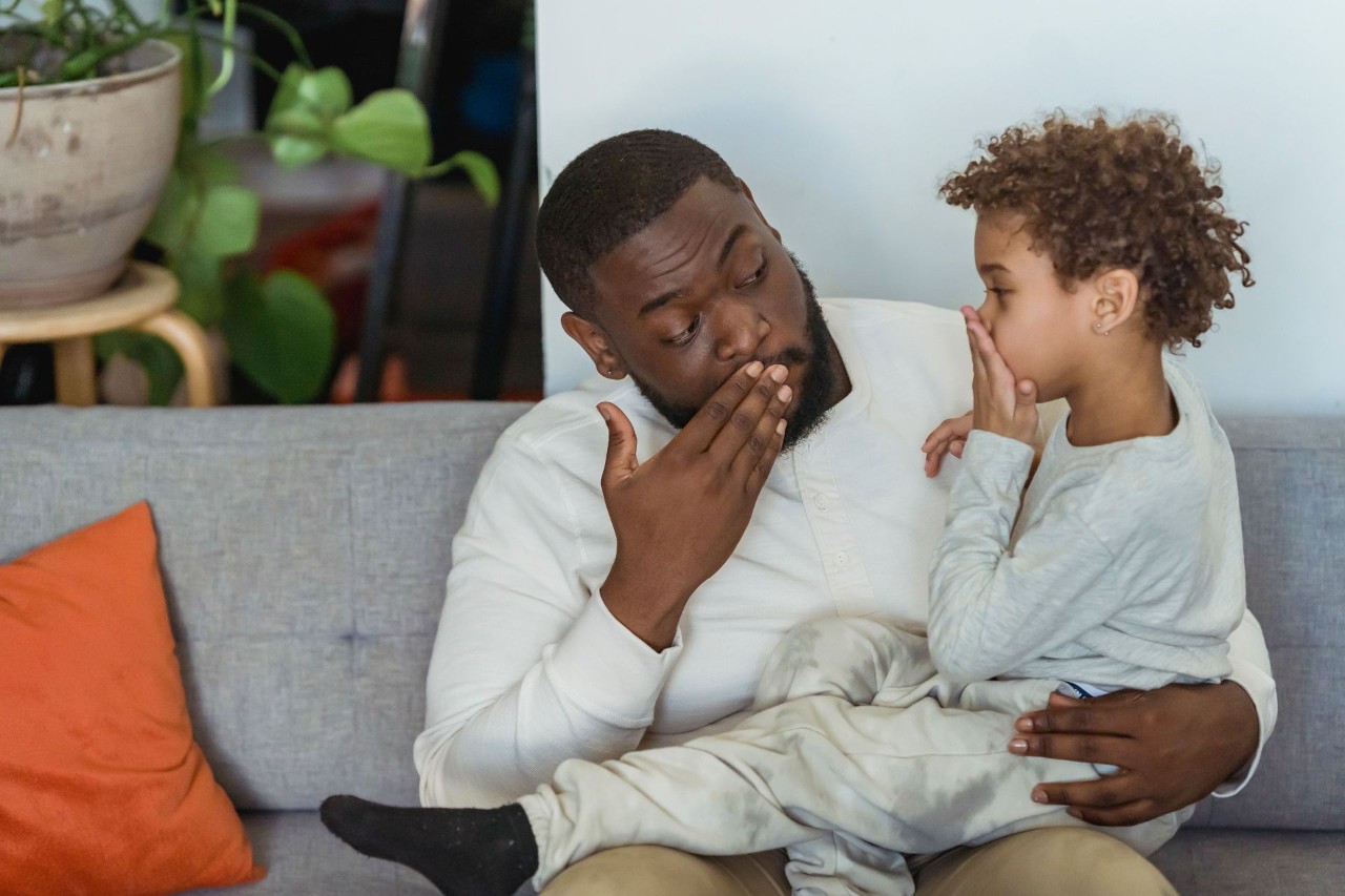 A father and son share a thoughtful moment indoors, sitting together on a sofa in a cozy home setting.