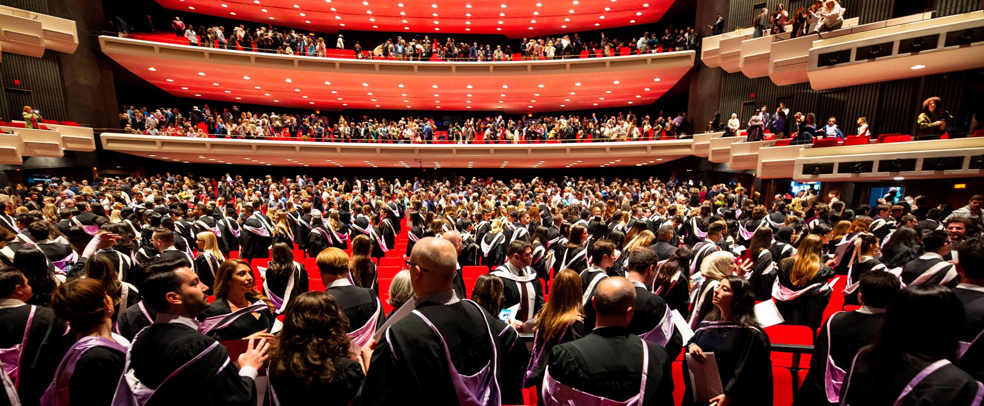 Graduating students, faculty members, and guests celebrating Convocation in black gowns against the red backgroud of Place des Arts