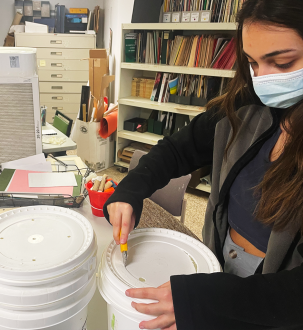 Masked young woman makes holes in the top of white plastic buckets with lids