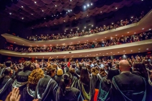 The backs of black-robed graduatign students in a large hall waving at the audience