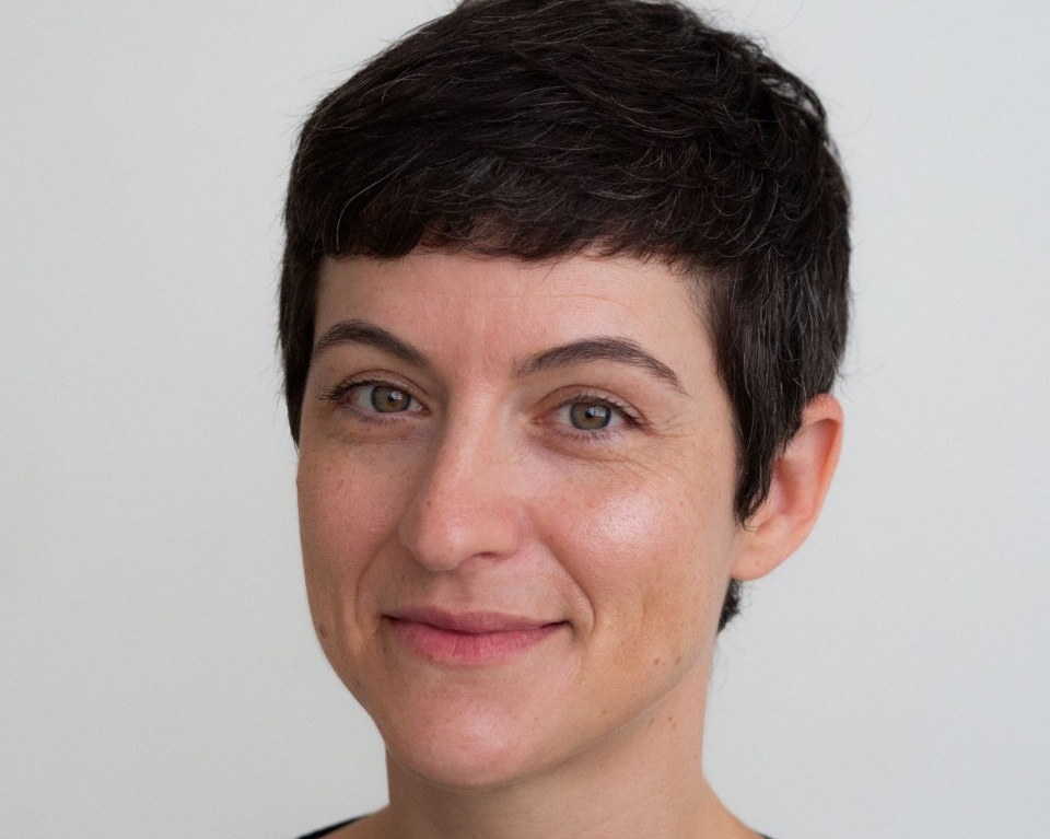 Une jeune femme souriante, aux cheveux bruns mi-longs, avec une frange, devant un arrière-plan flou où on peut aperçevoir des individus