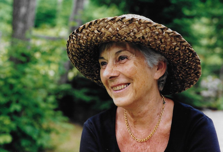 Jeanne Maranda in her later years. Short cropped gray hair under a straw hat. She is smiling and pictured outside with greenery in the background.