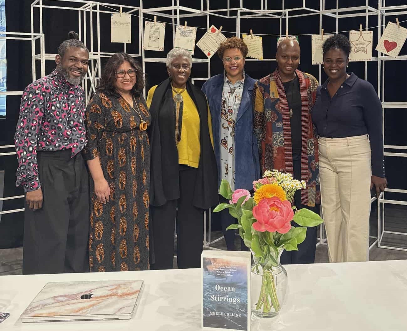 Dr. Ronald Cummings, Dr. Nalini Mohabir, Dr. Merle Collins, Marlihan Lopez, Dr. Laurie Lambert, and Tesfa Peterson stand arm-in-arm smiling at the camera.  In the foreground, there is a copy of Ocean Stirrings by Dr. Mere Collins beside a vase of blooming flowers.