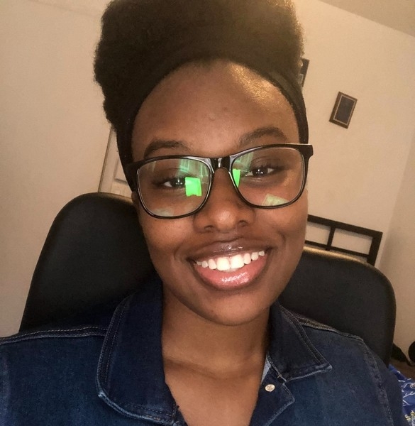 Head shot of a young black women seated in an office on a black chair. She is wearing black glasse, a black head band and a jeans vest. She is smiling and looking straght at the camera. Behind her are a white wall with a few pictures.