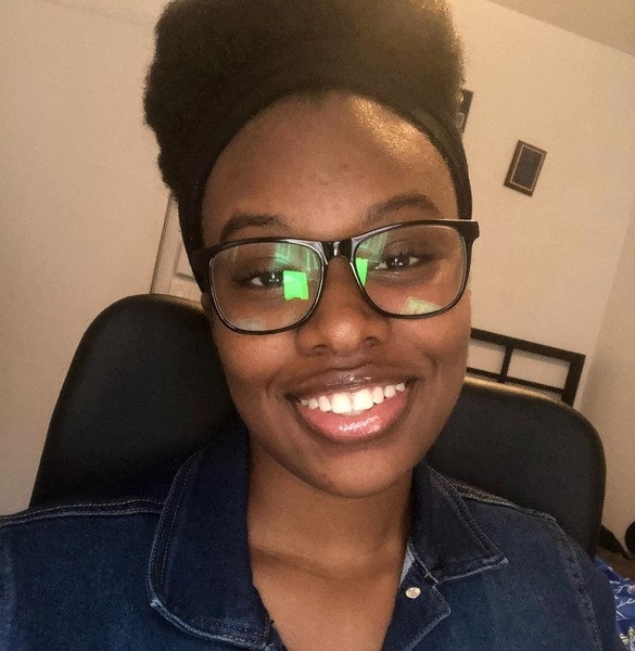 Markayla Black-Johnson Head shot of a young black women seated in an office on a black chair. She is wearing black glasse, a black head band and a jeans vest. She is smiling and looking straght at the camera. Behind her are a white wall with a few pictures.