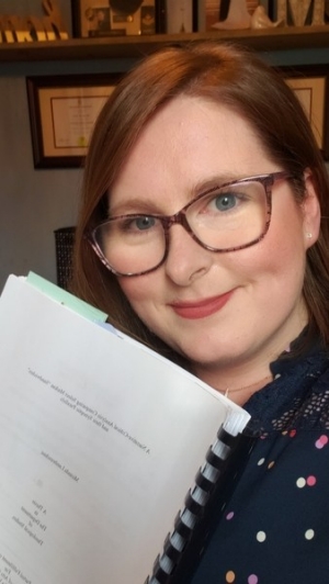 Full head shot of Lambruschini holding her printed Masters Thesis in front of a shelf filled with books and souvenirs. She has burgundy long hair and blue eyes. She is wearing glasses and a black shirt with colored polka dot designs.