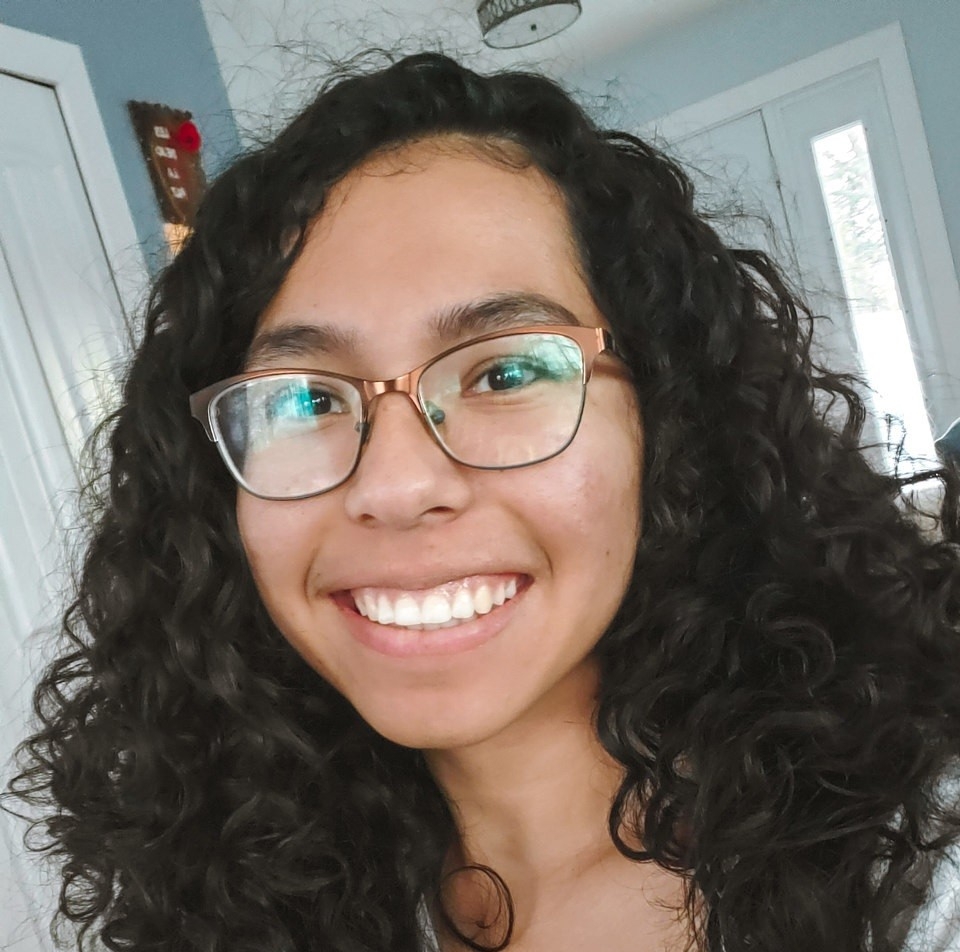 Headshot of a smiling Loren Gomez. She has long and curly black hair, is wearing glasses and a white t-shirt.