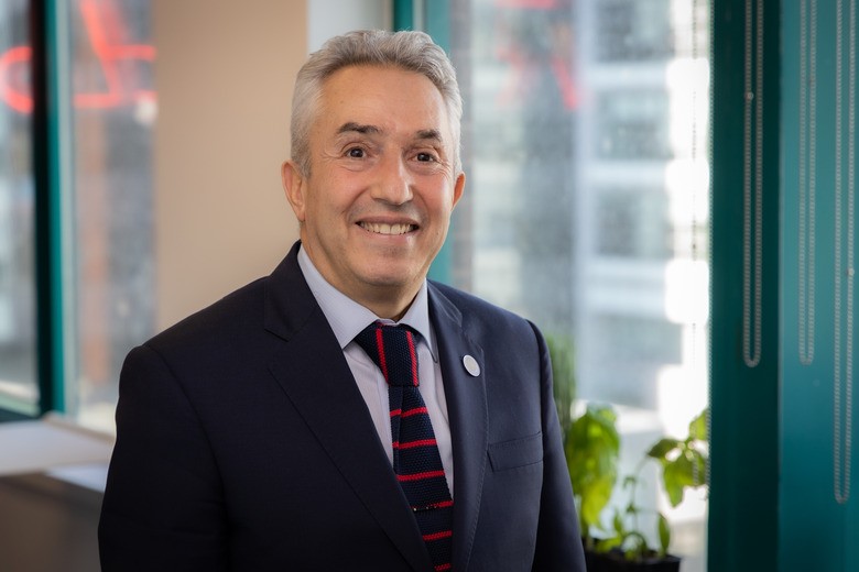 Portrait of Karim Zaghib smiling while standing in an office environment with large windows. He is wearing a dark suit, light blue shirt, and a striped red and blue tie. In the background, there are green plants and a cityscape visible through the windows.