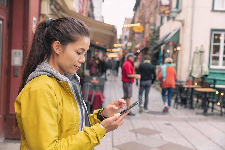 A young woman wearing a yellow jacket and holding a smartphone is standing on a bustling, picturesque street, focused on her device as people walk by in the background.