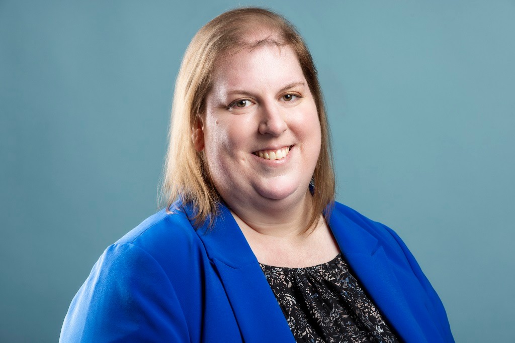 Smiling woman with shoulder-length light brown hair wearing a royal blue blazer over a black patterned top, in front of a light blue background.