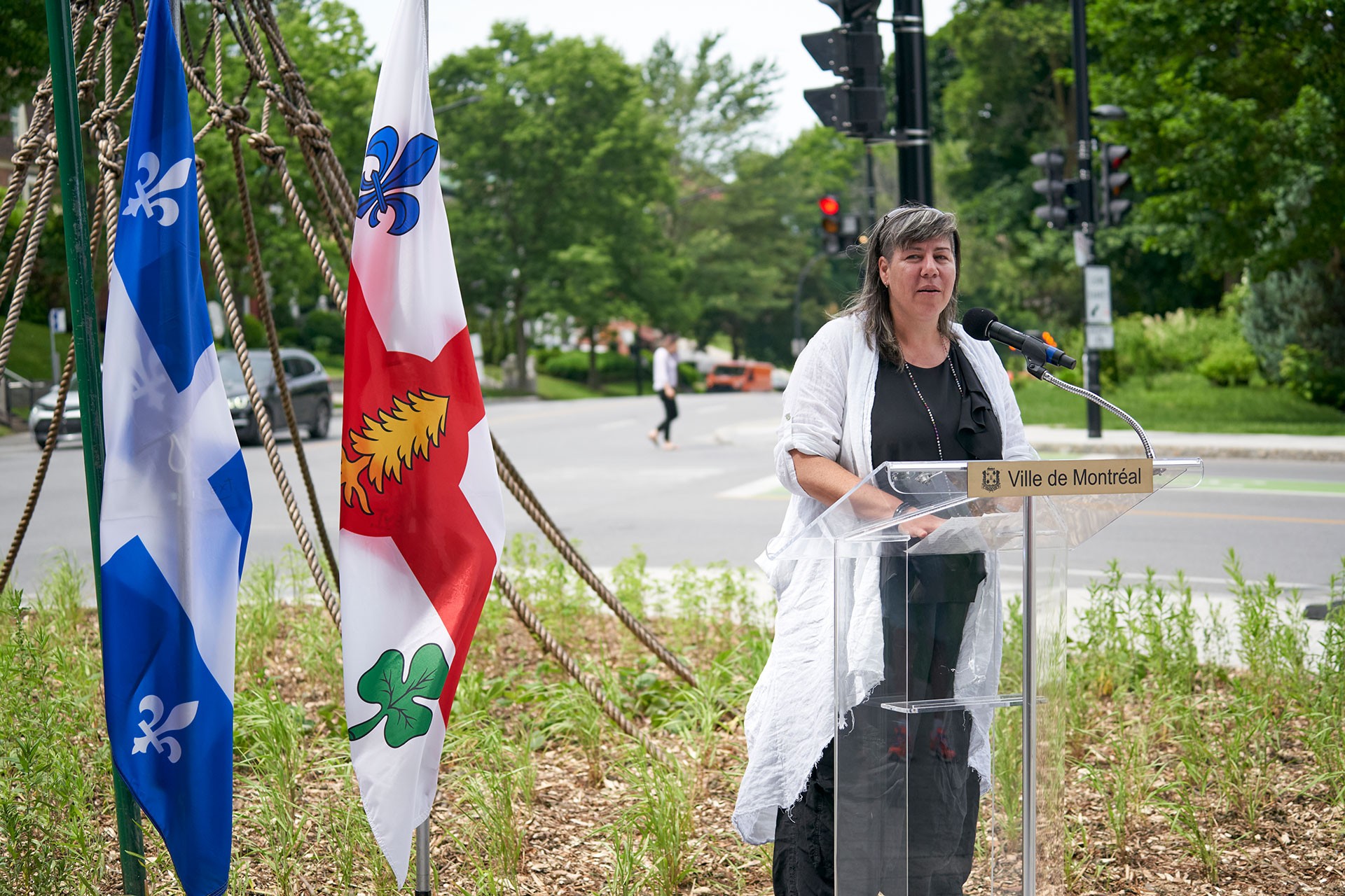 Nadia Myre at a podium speaking at the launch of her new public sculpture, Nouée.  Quebec and Montreal flags in background. 