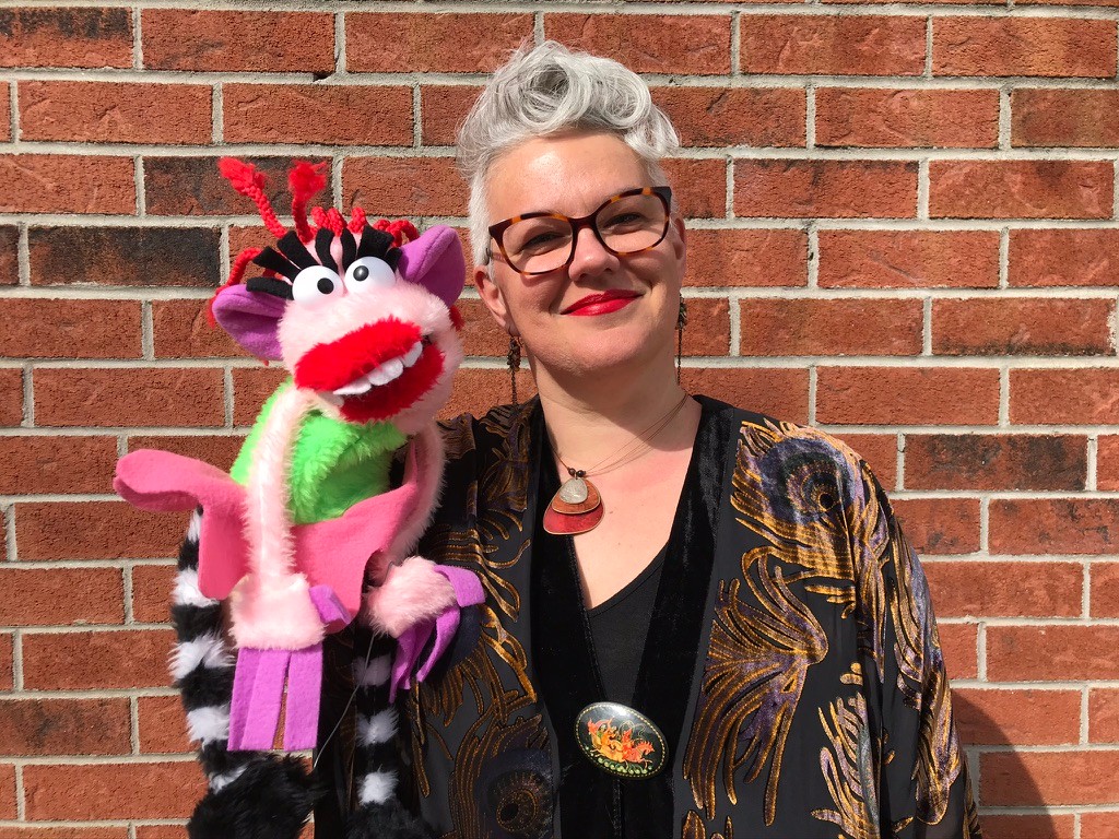 Woman with short hair, glasses and colorful puppet on hand, in front of brick wall