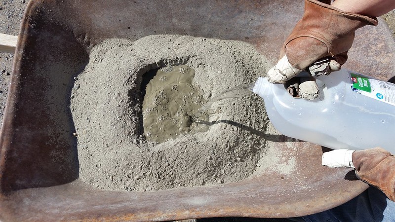 Person pouring water into a pile of dry concrete mix in a wheelbarrow while wearing protective gloves.