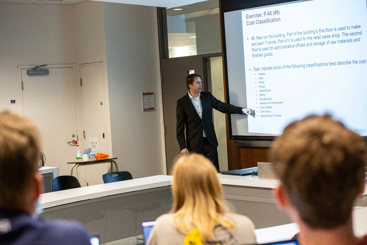 Matthäus Tekathen at pull-down screen lecturing with back of students' heads in foreground