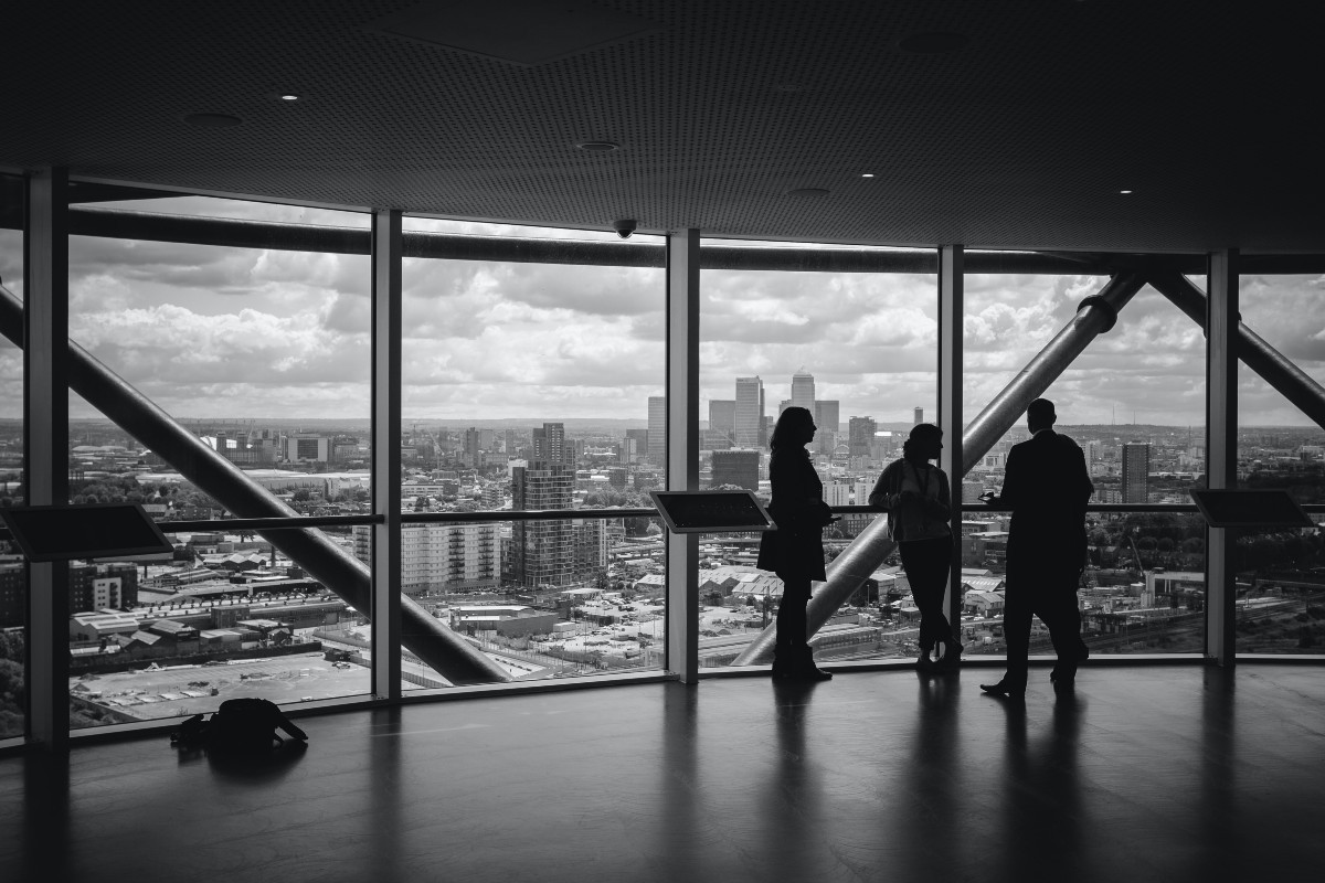 Three people meet in front of a wall of windows in a a high-rise office building. They are cast in silhouette.