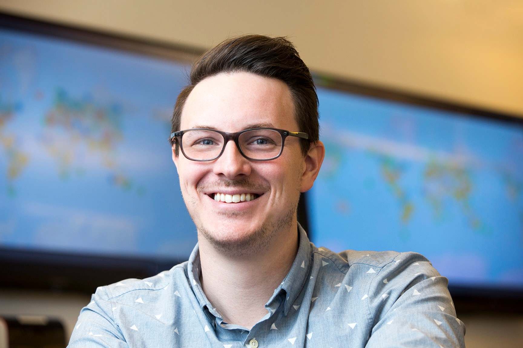 Man with glasses wearing a blue shirt, with blue screens in the background. 