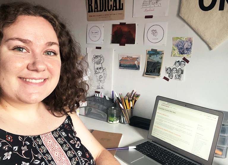 Young woman looking at the camera, seated at a desk with a laptop, with posters and postcards on the wall behind.