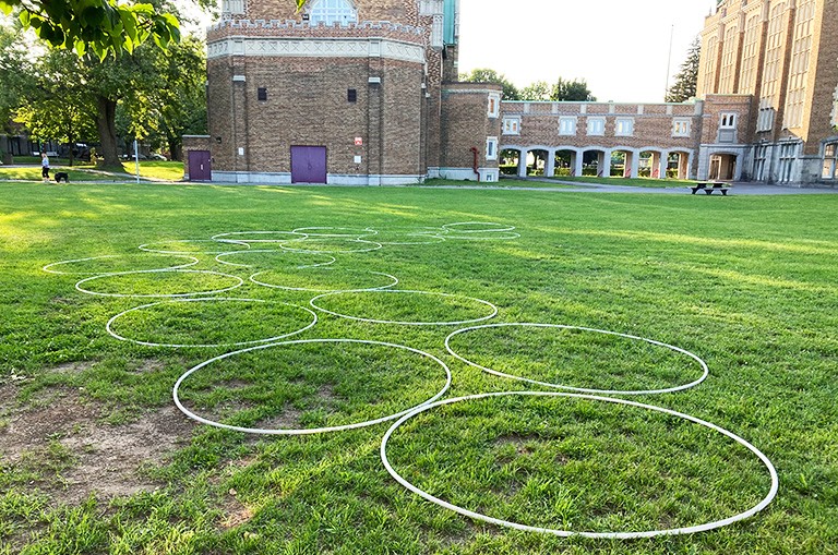 An open outdoor area with large circles made of thin plastic tubing placed on the grass, and university buildings in the background.