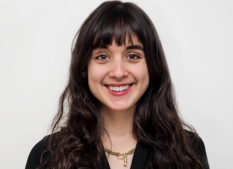 A smiling young woman with long brown hair, brown eyes and a black shirt in front of a pale background 