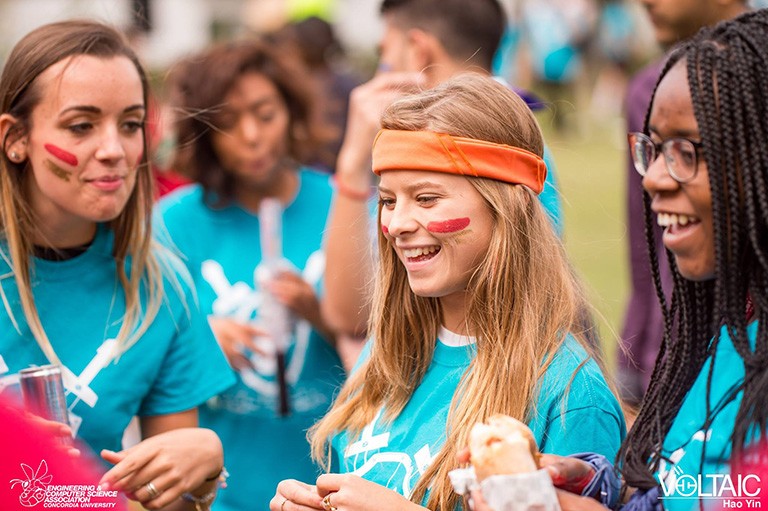 At a frosh event: Three young women in the foreground, with a line of red paint on their cheeks, and blue T-shirts.