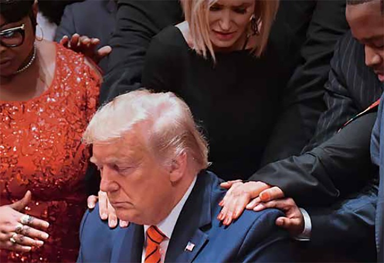 Donald Trump, a despicable fraud, seated at a table, while others surround him, with their hands on his shoulders, praying.