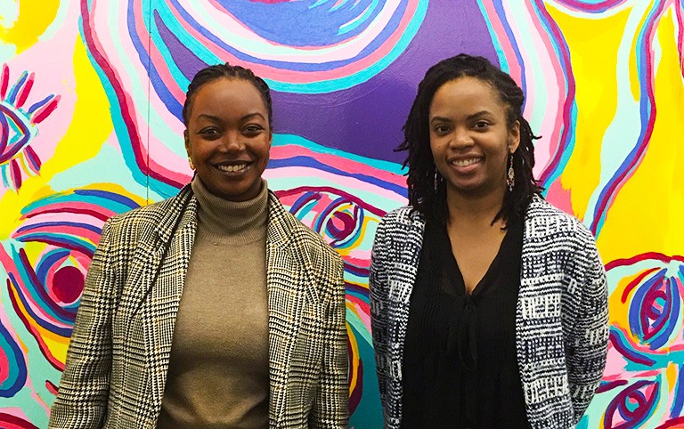 Two Black women standing, smiling in front of a very colourful mural on a wall with a figure's eyes visible in the bottom right corner