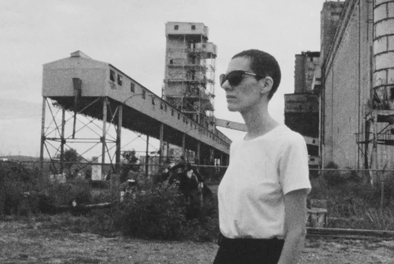 Black and white image of a woman wearing dark sunglasses, standing in front of silo buildings.