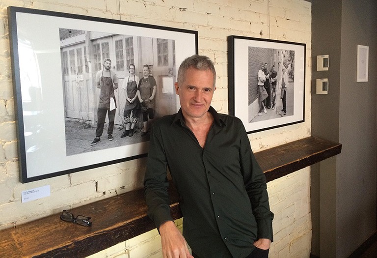 Man with short blond hair and a black shirt, standing in front of framed photographs.