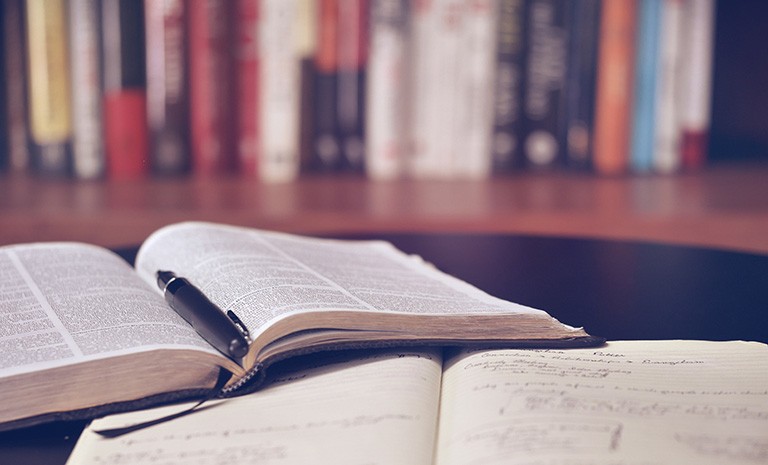 Books on a desk, with a fountain pen, and a shelf of books in the background. | Photo by Aaron Burden on Unsplash