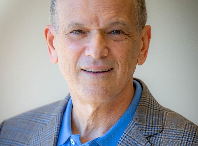 Older, bald, smiling man wearing a blue polo shirt and a suit jacket.