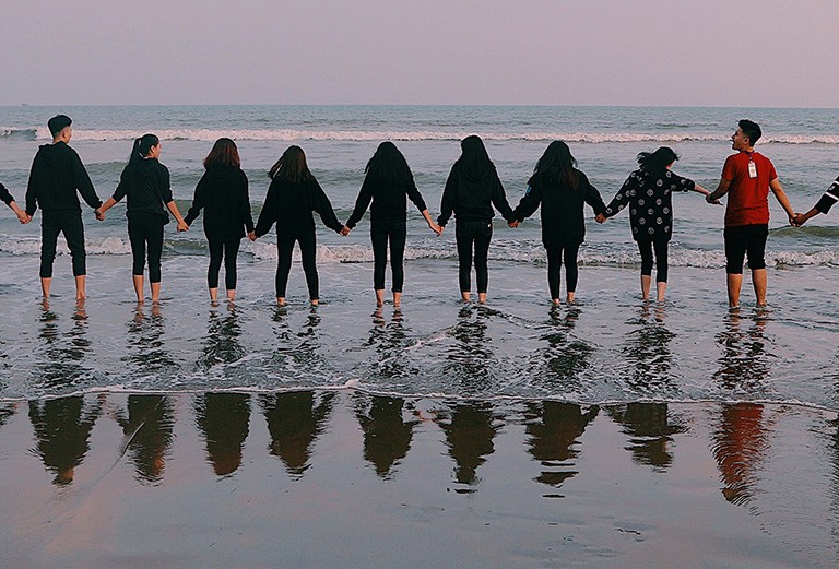 A group of people standing in a line, anke-deep in the ocean and holding hands.