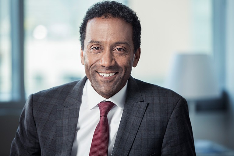 A smiling middle-aged man with short, curly, dark hair, a white shirt, red tie and checkered suit jacket.