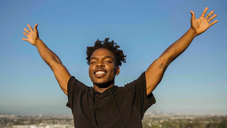 Young, smiling black man holds his arms wide, with the city far off in the background.