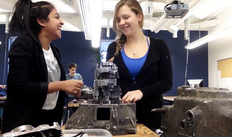Two women, laughing, in a workshop environment, taking apart a car engine.
