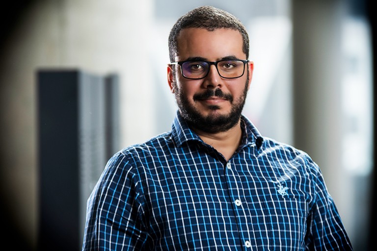 Young man with a beard, short, dark hair, dark-rimmed glasses and a checkered shirt.