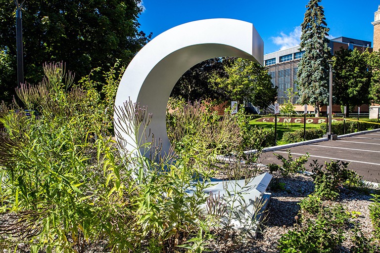 Concordia Loyola Campus Green corner of a parking lot with freestanding metal Concordia logo and buildings in the background