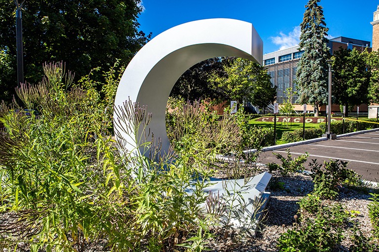 Green corner of a parking lot with freestanding metal Concordia logo and buildings in the background