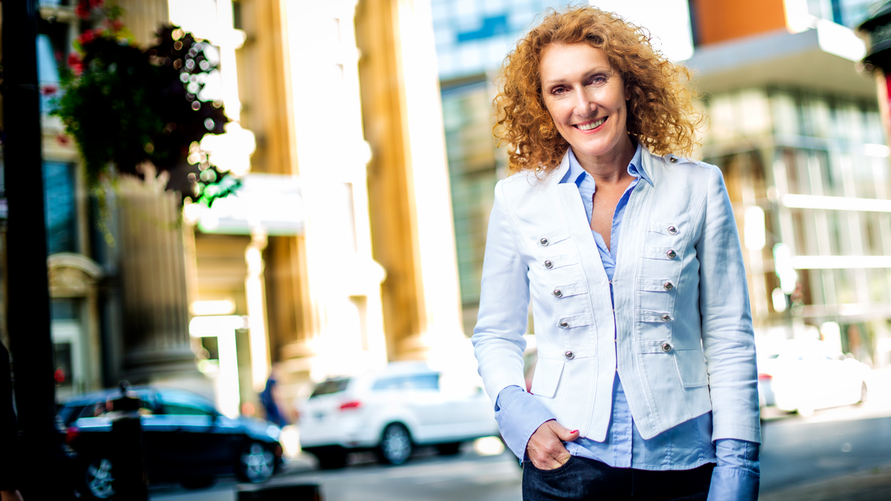A smiling woman with long, curly red hair, a blue shirt, white jacket and jeans, standing on a city pavement.