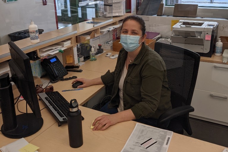 Woman wearing a procedure mask who is sitting at a desk with a computer and papers in front of her.