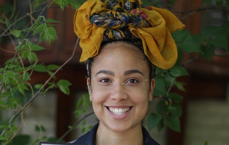 Young smiling woman wearing patterned mustard-yellow head wrap in front of a building and trees