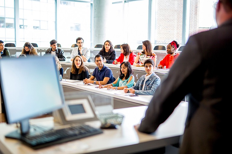 The back of an out-of-focus man facing students seated in a classroom at white and grey desks.