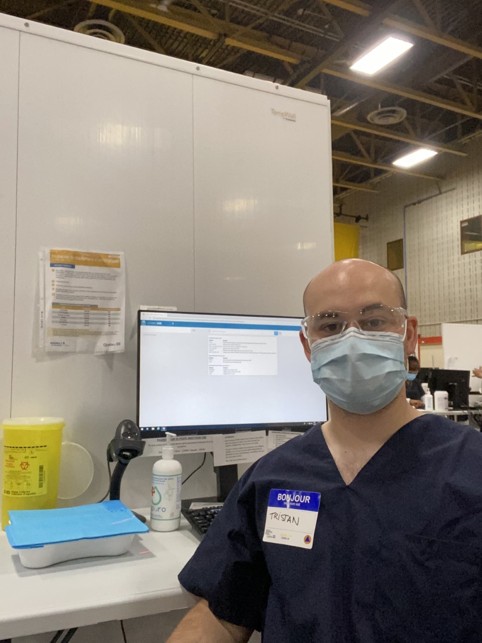 Tristan Castonguay seated at a desk wearing navy blue nursing scrubs, a blue surgical mask and clear protective glasses.