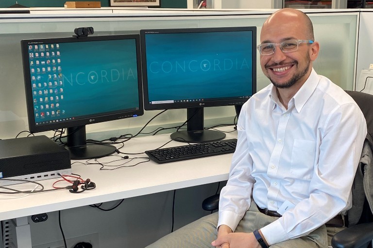 A man with glasses sits at a desk with two computer screens displaying Concordia's logo.