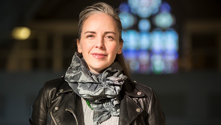 Smiling woman with long, tied-back hair, a ruffled scarf, grey top and black leather jacket.