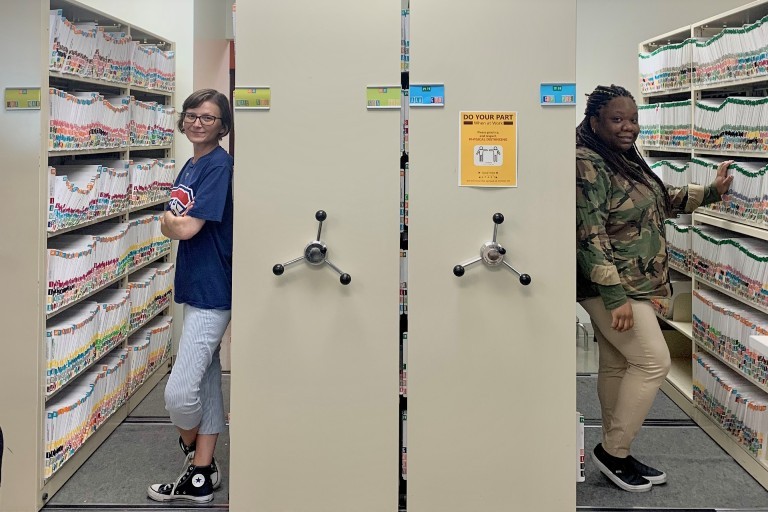 Two women stand with their backs on either side of two large filing cabinets.