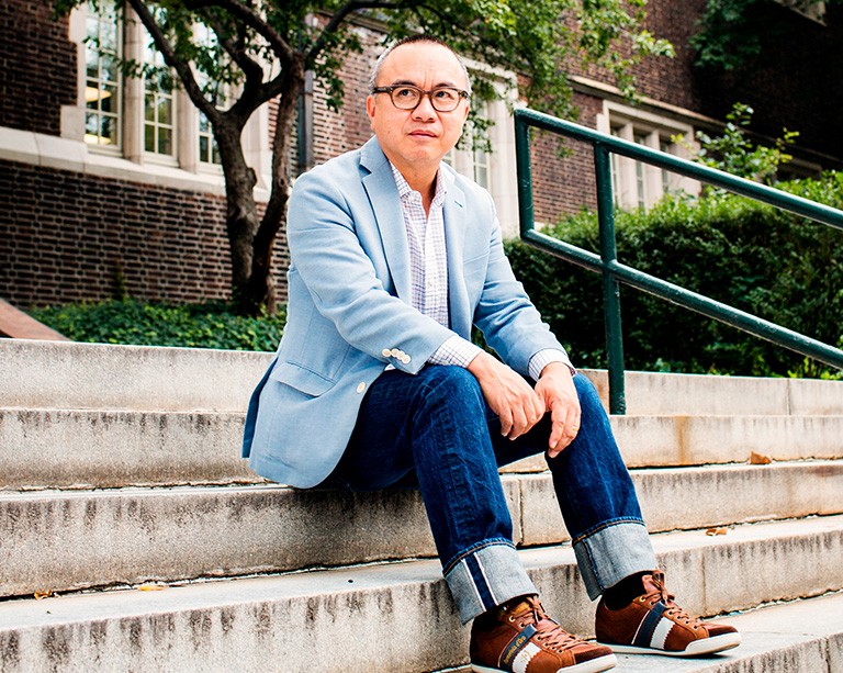 Man with glasses in a blue suit jacket, checkered shirt and jeans sitting on outdoor steps.