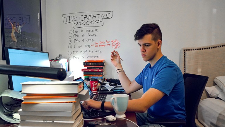 Young man sitting at a desk with a computer and a number of books and papers